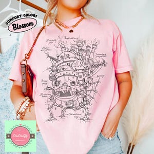 May include: A light pink t-shirt featuring a detailed black and white illustration of a mechanical structure with the text "COMFORT COLORS Blossom". The design includes architectural elements and technical annotations. The shirt is a comfortable fit.