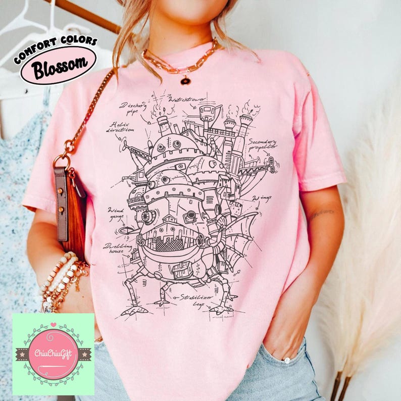 May include: A light pink t-shirt featuring a detailed black and white illustration of a mechanical structure with the text "COMFORT COLORS Blossom". The design includes architectural elements and technical annotations. The shirt is a comfortable fit.
