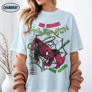 May include: A light blue t-shirt featuring a graphic of the comic book character Spider-Man in red and black, surrounded by a black web design. The text reads THE AMAZING SPIDER-MAN and QUEENS NEW YORK in green and red lettering on the front.