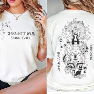May include: Cream-colored t-shirt featuring a black and white illustration of Studio Ghibli characters. The front displays a Totoro design with Japanese text and "STUDIO GHIBLI." The back showcases a detailed graphic of various characters and the text "STUDIO GHIBLI."