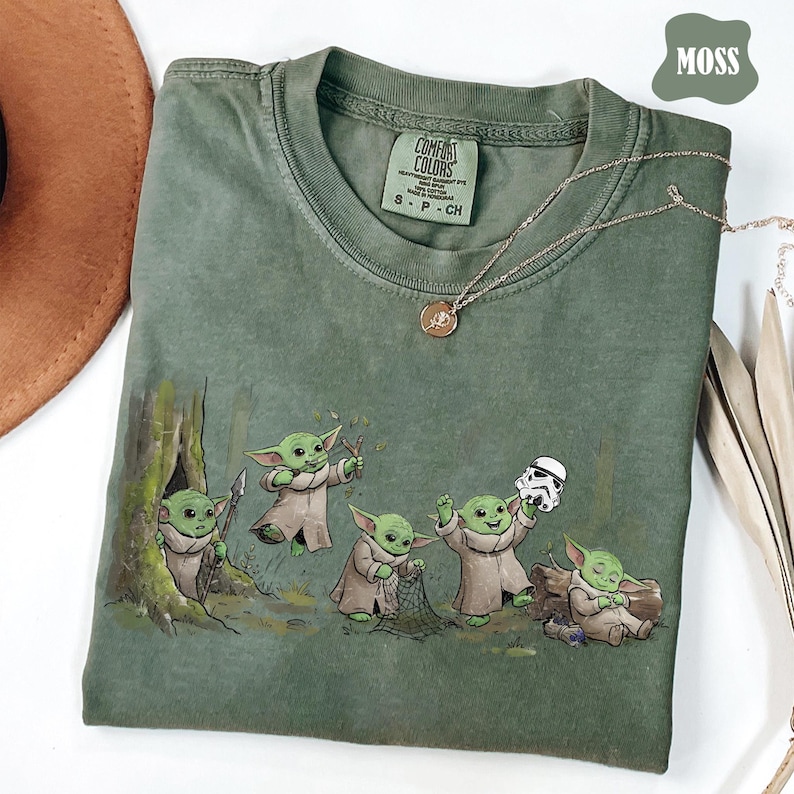以下が含まれることがあります： モスグリーンのクルーネックTシャツ。ローブを着た5体の小さな緑色の生き物のグラフィックプリント入り。槍、パチンコ、網、白いヘルメットを持つものや、丸太に座っているものが描かれている。首元にはComfort Colorsのラベル付き。