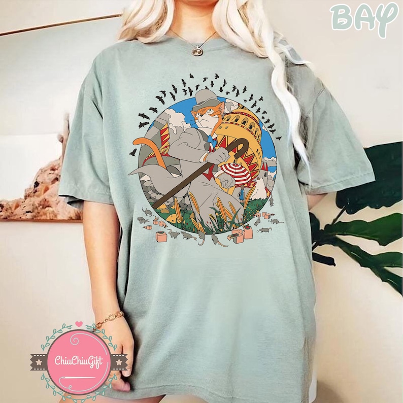 May include: A light green t-shirt featuring a circular graphic of a cat in a suit with a cane, surrounded by bats and a building. The graphic has a cartoon style with a vintage aesthetic. The shirt has a relaxed fit.