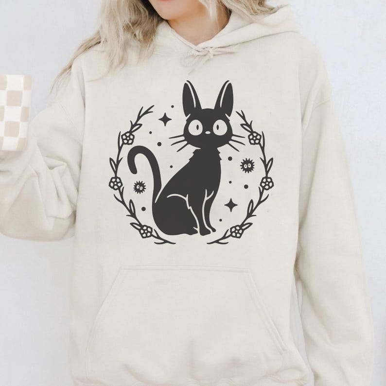 May include: Cream-colored hoodie featuring a black cat illustration within a floral wreath. The cat has large, round eyes and is surrounded by stars and floral accents. The hoodie has a drawstring hood and a front pocket.