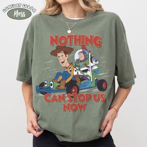 May include: A moss green short-sleeve t-shirt featuring a graphic of Woody and Buzz Lightyear riding a go-kart. The text above and below the characters reads NOTHING CAN STOP US NOW. A small oval label in the top left corner says COMFORT COLOR MOSS.