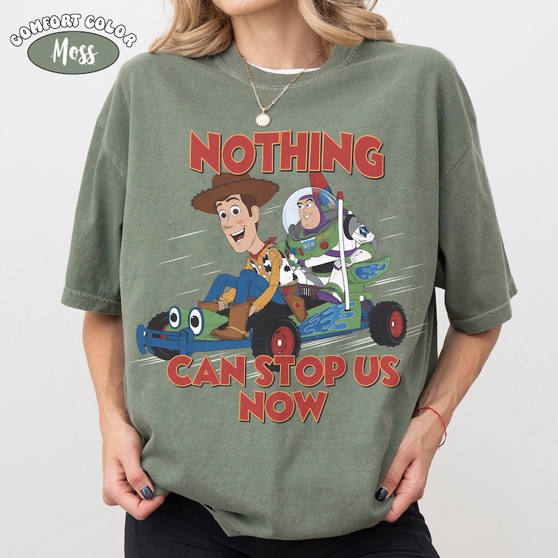 May include: A moss green short-sleeve t-shirt featuring a graphic of Woody and Buzz Lightyear riding a go-kart. The text above and below the characters reads NOTHING CAN STOP US NOW. A small oval label in the top left corner says COMFORT COLOR MOSS.