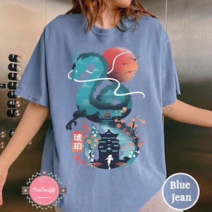 Puede incluir: Camiseta azul vaquero con un dise&ntilde;o de drag&oacute;n turquesa, un sol rojo y p&aacute;jaros volando. El drag&oacute;n envuelve un paisaje con una pagoda, flores de cerezo y el texto "琥珀". Una l&iacute;nea blanca curva alrededor del drag&oacute;n. La camiseta tiene un corte holgado.