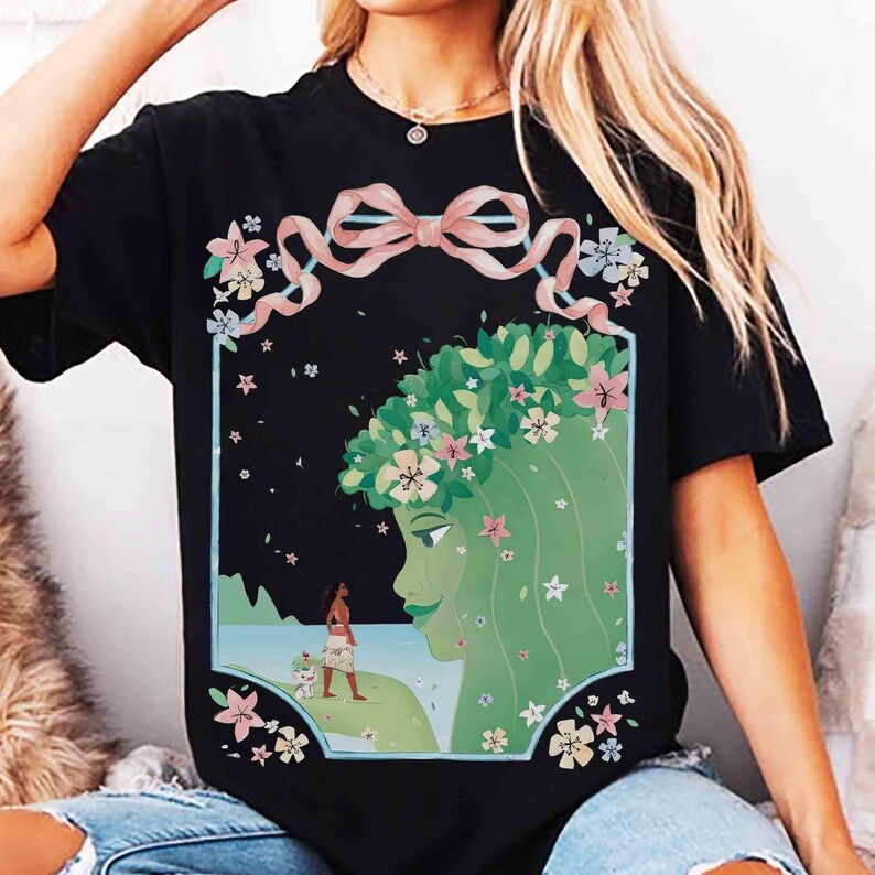 May include: Black t-shirt featuring a vibrant illustration of a green-skinned woman with a floral crown and a pink bow. The design includes a figure on a beach with a dog, surrounded by flowers.