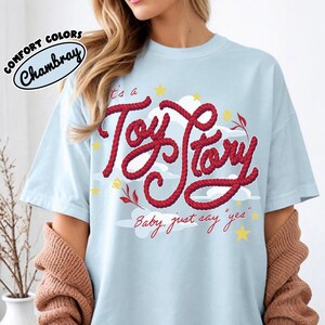 May include: A light blue t-shirt featuring a graphic print with the red rope-style text "It's a Toy Story" and "Baby, just say "yes"" in red cursive. The design includes white clouds, yellow stars, and small floral accents on the front.