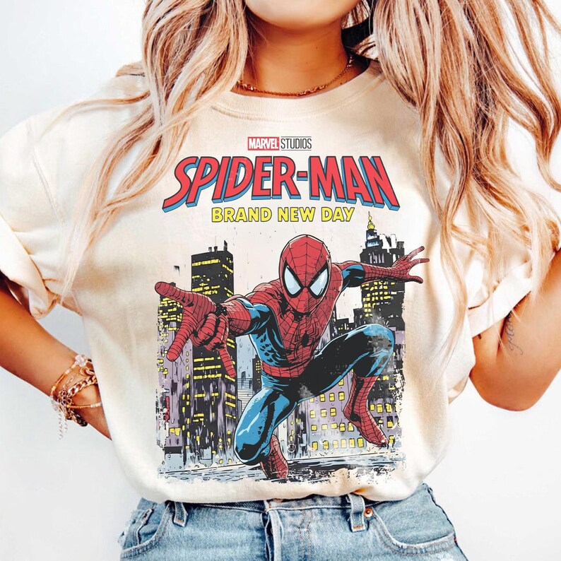 May include: Cream-colored t-shirt featuring a Marvel Studios Spider-Man graphic. The design includes the words "SPIDER-MAN" in red and blue, with "BRAND NEW DAY" below. The graphic depicts Spider-Man in a dynamic pose against a cityscape.