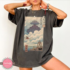 May include: Dark gray t-shirt featuring a Japanese anime-style illustration. The artwork depicts a floating castle, a flying machine, and a robot. Japanese text is visible on the design. The shirt has a relaxed fit.