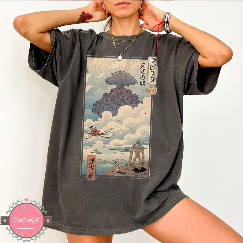 May include: Dark gray t-shirt featuring a Japanese anime-style illustration. The artwork depicts a floating castle, a flying machine, and a robot. Japanese text is visible on the design. The shirt has a relaxed fit.
