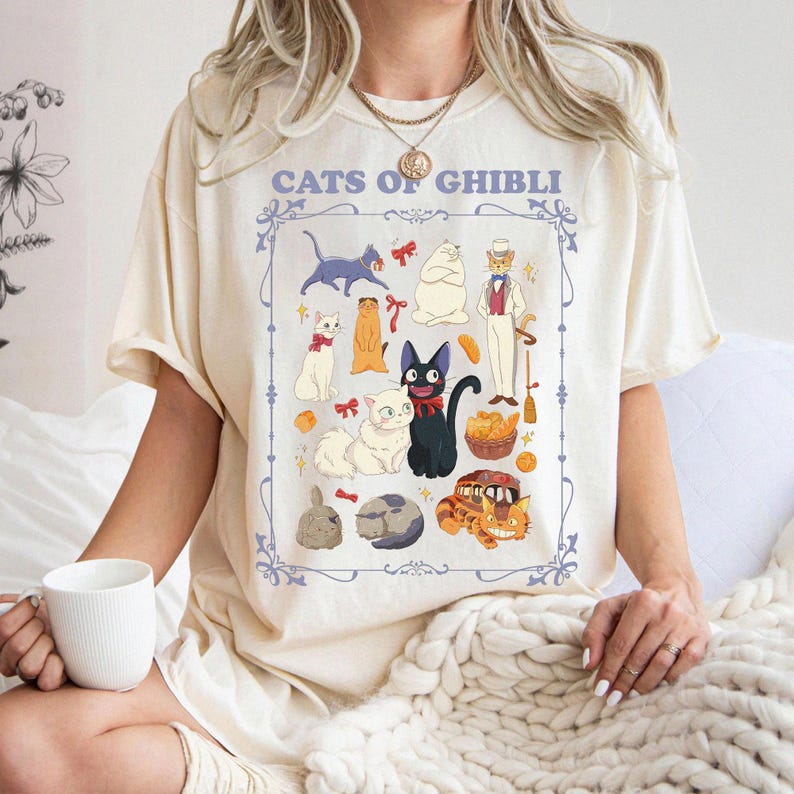 K&ouml;nnte beinhalten: Cremefarbenes T-Shirt mit einer Grafik verschiedener Zeichentrickkatzen und dem Text "CATS OF GHIBLI" in einem hellblauen Rahmen. Das Design zeigt mehrere Katzenfiguren in verschiedenen Posen und Farben.