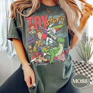 May include: A dark green t-shirt featuring a graphic print of Toy Story characters including Woody, Buzz Lightyear, Jessie, Rex, Slinky Dog, Bullseye, and Hamm. The text reads Toy Story, since 1995, and identifies each character by their name.
