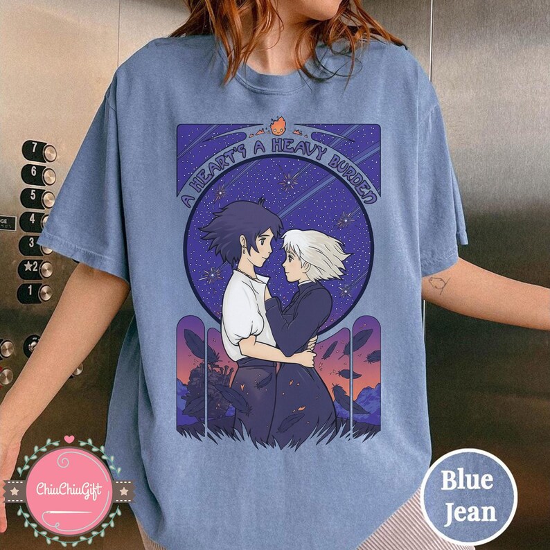 Puede incluir: Camiseta azul vaquera con un dise&ntilde;o de la pel&iacute;cula de animaci&oacute;n, El castillo ambulante. El dise&ntilde;o muestra a dos personajes abraz&aacute;ndose, sobre un cielo estrellado, con el texto "A HEART'S A HEAVY BURDEN".