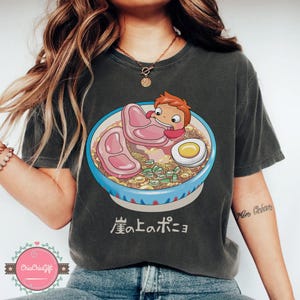 May include: Dark gray t-shirt featuring a colorful illustration of a bowl of ramen with a cartoon character. The ramen includes noodles, meat, an egg, and green onions. Japanese text is below the bowl.