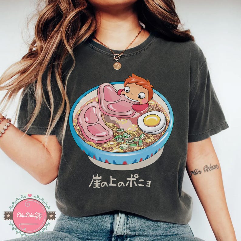 May include: Dark gray t-shirt featuring a colorful illustration of a bowl of ramen with a cartoon character. The ramen includes noodles, meat, an egg, and green onions. Japanese text is below the bowl.
