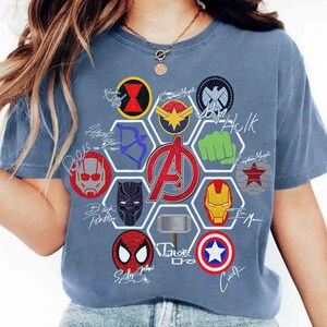 May include: A dusty blue t-shirt featuring a large central Avengers logo surrounded by a hexagonal pattern of various superhero icons and symbols, including masks, weapons, and emblems, accompanied by handwritten-style character names in white ink.