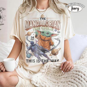 May include: An ivory t-shirt featuring a graphic print of The Mandalorian and The Child in space. The design includes planets, stars, and text reading THE MANDALORIAN THE CHILD 24 JULY 12c THIS IS THE WAY. A small circular label in the corner says COMFORT COLOR IVORY.