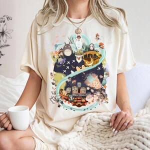 May include: Cream-colored t-shirt featuring a colorful graphic design. The design includes various cartoon characters and elements, set against a dark blue and green background. The shirt has short sleeves and a relaxed fit.
