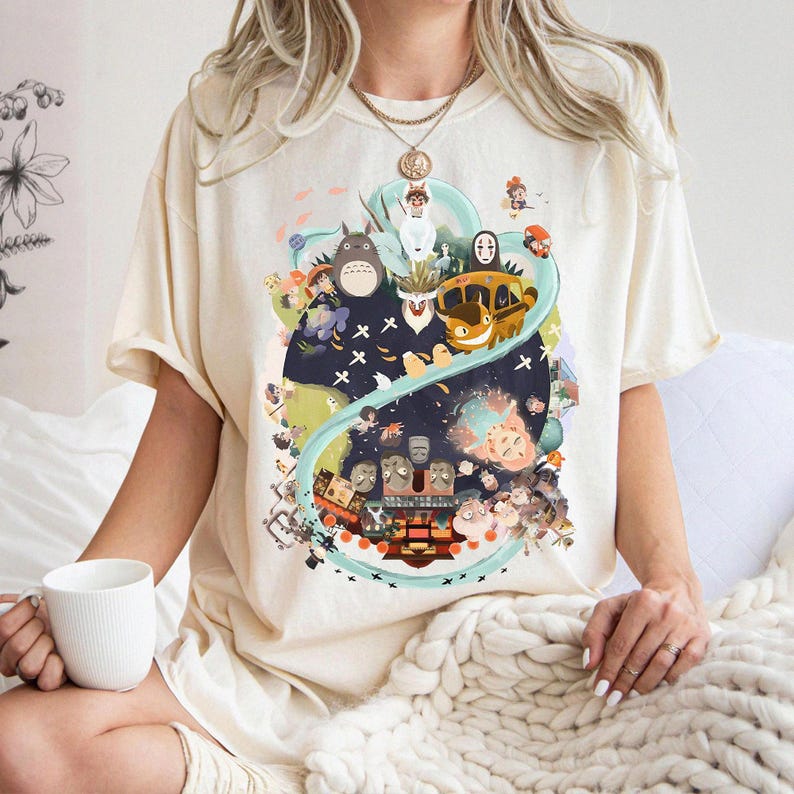 May include: Cream-colored t-shirt featuring a colorful graphic design. The design includes various cartoon characters and elements, set against a dark blue and green background. The shirt has short sleeves and a relaxed fit.