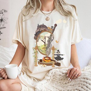 Op de afbeelding: Cr&egrave;mekleurig T-shirt met een grillig klokontwerp met tandwielen, een huis en een figuur met hoge hoed. De wijzerplaat heeft zwarte wijzers. Het ontwerp bevat een klein huis, een maan en een stoom effect.