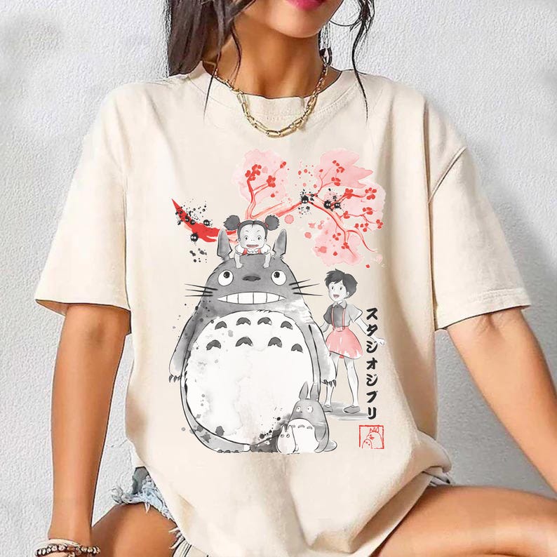 Pode incluir: Camiseta creme com uma ilustra&ccedil;&atilde;o em aquarela de personagens do filme de anima&ccedil;&atilde;o "Meu Vizinho Totoro". O design inclui Totoro, uma menina e uma pequena criatura semelhante a um coelho sob um galho de flor de cerejeira rosa. Tamb&eacute;m h&aacute; texto japon&ecirc;s.