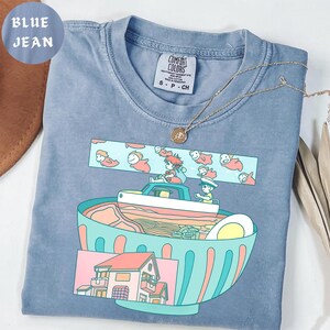 Puede incluir: Una camiseta azul vaquero Comfort Colors con un gr&aacute;fico colorido de un cuenco de ramen, un barco con personajes y una casa. La camiseta tiene una etiqueta peque&ntilde;a y un collar dorado. El texto "BLUE JEAN" est&aacute; en un c&iacute;rculo en la esquina superior izquierda.