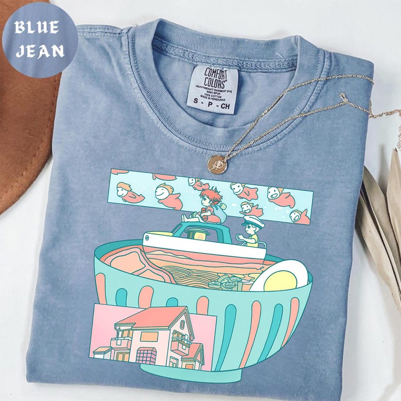 Puede incluir: Una camiseta azul vaquero Comfort Colors con un gr&aacute;fico colorido de un cuenco de ramen, un barco con personajes y una casa. La camiseta tiene una etiqueta peque&ntilde;a y un collar dorado. El texto "BLUE JEAN" est&aacute; en un c&iacute;rculo en la esquina superior izquierda.