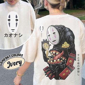 May include: Ivory-colored t-shirt featuring a graphic of a character from the animated film "Spirited Away." The front displays a white mask with black details and Japanese text. The back showcases a larger illustration of the character with additional Japanese text and red accents.