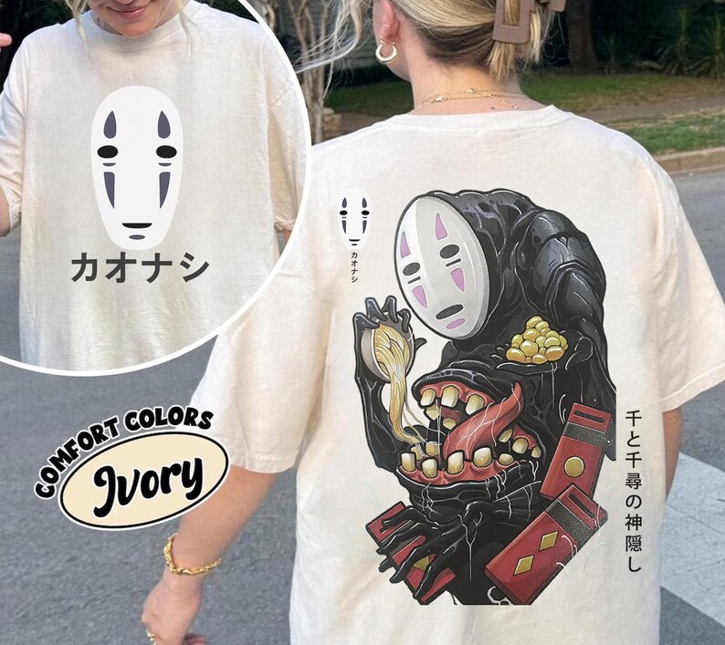 May include: Ivory-colored t-shirt featuring a graphic of a character from the animated film "Spirited Away." The front displays a white mask with black details and Japanese text. The back showcases a larger illustration of the character with additional Japanese text and red accents.