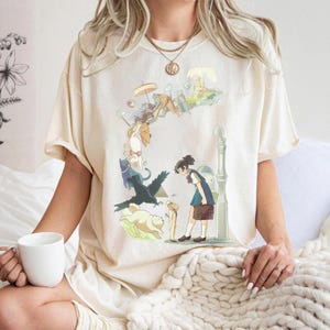 Puede incluir: Camiseta color crema con una ilustraci&oacute;n caprichosa de estilo anime. El dise&ntilde;o incluye una ni&ntilde;a con uniforme escolar, gatos, un cuervo y otros elementos de fantas&iacute;a. La camiseta tiene mangas cortas y un corte holgado.
