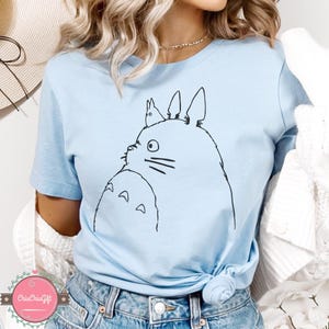 Puede incluir: Camiseta azul claro con un dibujo lineal negro de un personaje de Totoro y un peque&ntilde;o conejo. El dise&ntilde;o est&aacute; centrado en la camiseta. La camiseta est&aacute; hecha de un material suave.