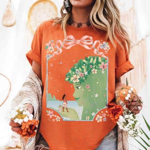 May include: Orange t-shirt featuring a floral design with a woman's profile and a smaller figure on a beach. The design includes a green-haired woman with a flower crown, a pink bow, and floral accents. The shirt has rolled-up sleeves.