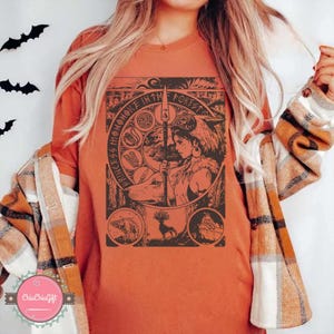 Puede incluir: Camiseta naranja con un gr&aacute;fico negro y marr&oacute;n de una mujer, un lobo, un ciervo y un oso, con el texto "PRINCESS MONONOKE IN THE FOREST". La camiseta es de color naranja liso.