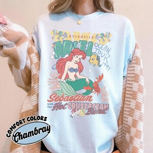 Op de afbeelding: Een lichtblauw sweatshirt met ronde hals en een grafische print van Ariel, Botje en Sebastiaan uit De Kleine Zeemeermin. De tekst luidt Atlantica Ariel Ocean Tour 1991, Sebastian with the Hot Crustacean Band. Een beige en wit geblokt vest is eroverheen gedragen.
