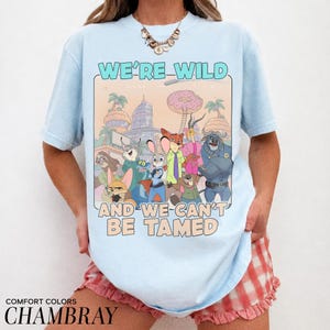 May include: A light blue short-sleeve t-shirt featuring a graphic print of various animated animal characters from Zootopia. The text WE'RE WILD AND WE CAN'T BE TAMED is printed in large letters above and below the colorful illustration.