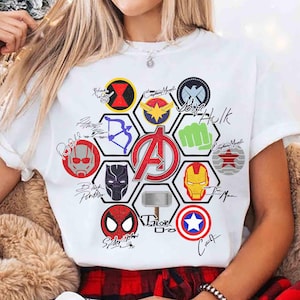 May include: A white t-shirt featuring a central red Avengers logo surrounded by a hexagonal pattern of various superhero icons, including masks, symbols, and weapons, accompanied by handwritten-style character names in black ink.