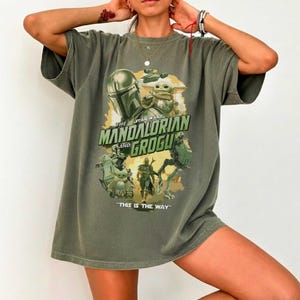 May include: An olive green t-shirt featuring a graphic print of The Mandalorian and Grogu. The design includes the text "THE MANDALORIAN AND GROGU" and "THIS IS THE WAY" in green lettering, surrounded by various illustrations of the characters.