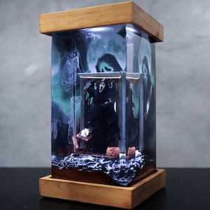 Puede incluir: Una vitrina rectangular con marco de madera que presenta una escena de terror. En el interior, una figura negra de Ghostface se alza frente a un fondo de luna llena y efectos de humo. La base y la parte superior son de madera clara.