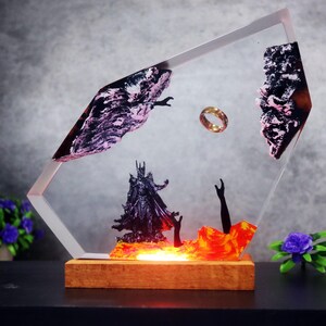 Può includere: Scultura decorativa in resina raffigurante una scena con un anello d'oro, una figura oscura ed elementi infuocati. L'opera è racchiusa in una cornice trasparente e angolata, posta su una base di legno.