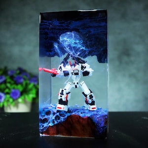 Könnte beinhalten: Eine weiße und schwarze Roboter-Actionfigur mit roten Akzenten, die eine rote Waffe hält und in einem transparenten Harzblock eingeschlossen ist. Das Harz weist ein blaues, wirbelndes Design auf, und die Figur steht auf einer braunen und blauen Basis.