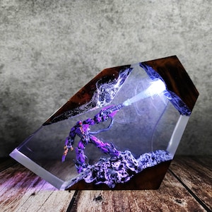 May include: A purple and black action figure encased in clear resin with a wooden frame. The figure is posed with a weapon, emitting a blue light. The resin block sits on a wooden surface.