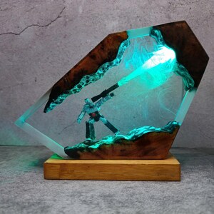 May include: A unique resin art piece featuring a robot figure with a glowing green beam. The artwork is encased in a clear resin with wooden accents and a wooden base. The piece has a futuristic, sci-fi theme.