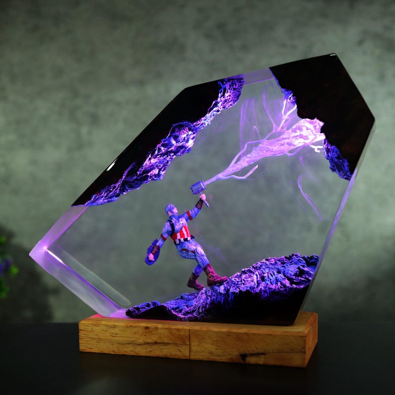 K&ouml;nnte beinhalten: Dekorative Resin-Skulptur mit Captain America mit Schild und Hammer, vor einer lila-schwarzen Landschaft. Die Skulptur wird mit lila Licht beleuchtet und auf einem Holzsockel montiert.