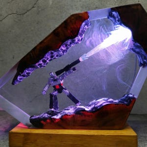 May include: A unique resin art piece featuring a silver robot figure with a black weapon, set within a clear, geometric frame. The frame has a wood-grain border with purple and red accents, and a bright light beam. The base is made of wood.