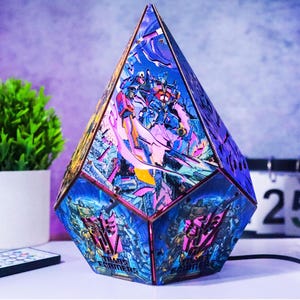 Könnte beinhalten: Eine geometrische Prismenlampe mit Transformers-Charakteren in Blau-, Rosa- und Lilatönen. Die Lampe hat ein facettenreiches Design, wobei das Transformers-Logo auf der Basis sichtbar ist. Die Lampe ist eingesteckt und leuchtet.