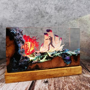 May include: A rectangular resin art piece featuring a miniature figure running towards a red, glowing structure. The base is wood, and the resin contains colorful, textured elements. The piece is illuminated from within, creating a dramatic effect.