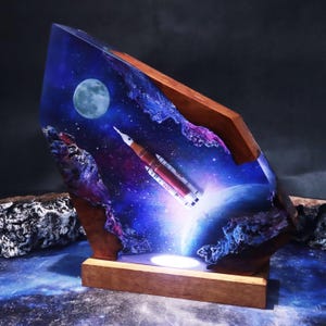 Könnte beinhalten: Ein einzigartiges Kunstwerk zum Thema Weltraum, das eine Rakete zeigt, die in eine lebendige Galaxie startet. Das Kunstwerk ist in klarem Harz eingeschlossen und hat einen Holzsockel und -rahmen. Die Szene umfasst einen Vollmond, Planeten und kosmische Elemente.
