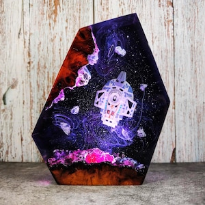 Könnte beinhalten: Ein sechseckiges Kunstwerk aus Kunstharz, das eine Weltraumszene mit einem Raumschiff und Himmelselementen darstellt. Das Kunstwerk verwendet Farbtöne von Lila, Orange und Schwarz, mit einem detaillierten Raumschiffdesign. Das Stück ist vor einem Holzgrund platziert.