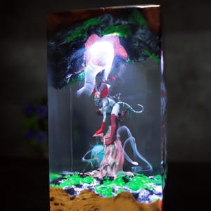 Könnte beinhalten: Harzskulptur mit einer Fantasy-Figur mit rotem Kopfschmuck und Stiefeln, die auf einer baumartigen Struktur posiert. Die Skulptur ist in einem transparenten Block mit einer leuchtenden Lichtquelle eingeschlossen, umgeben von grünen und braunen Elementen.
